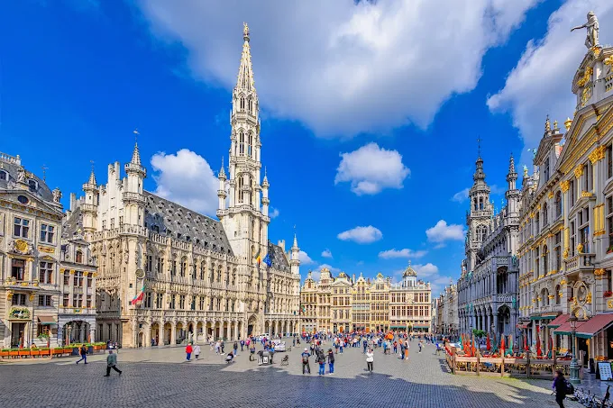 La Grand-Place de Bruxelles par temps clair : l’hôtel de ville et sa flèche gothique, les maisons de corporations et les passants sur les pavés.